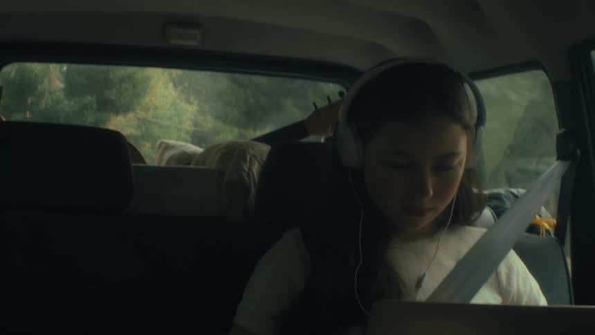 A young girl sits in the backseat of a moving car wearing large headphones and looking down at a laptop screen while a guitar rests in the trunk area behind her