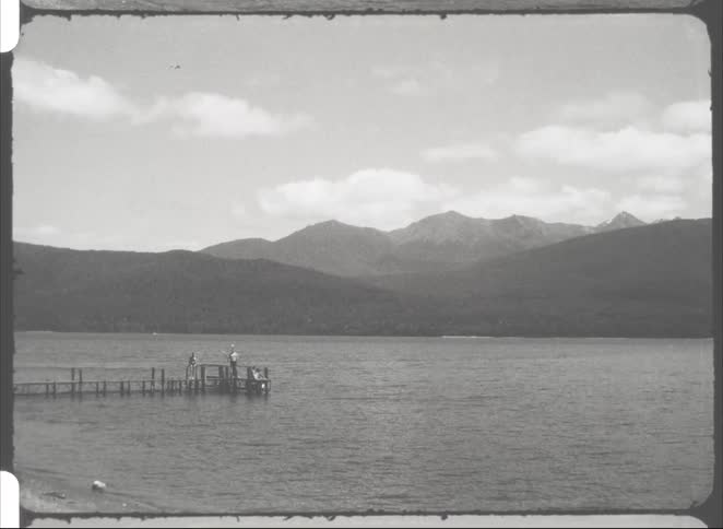Two people stand on the end of a long wooden pier extending into a vast lake waving their arms toward the camera while mountains and a dense forest line the horizon under a bright cloudy sky