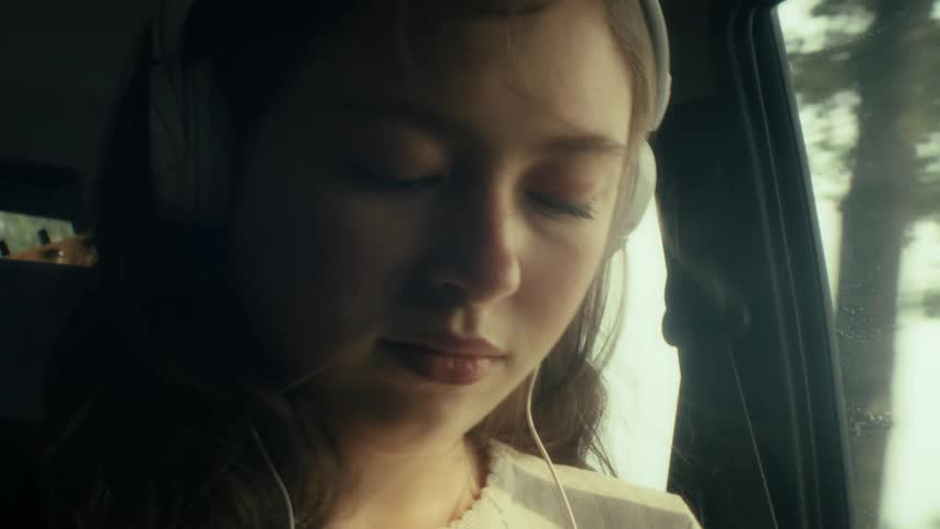 Teenage girl wears headphones as she sits in the back seat of an automobile