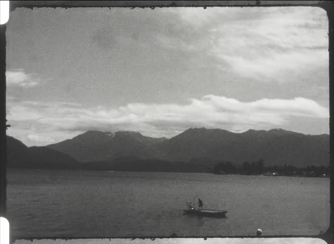 A person stands on a small boat in a vast body of water before a mountain range under a cloudy sky looking forward with a serene mood