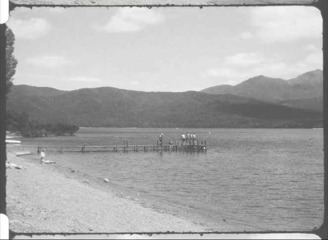 A group of people stands on a dock extending into the lake at midday while others gather along the nearby bank across the water a mountain range borders the far side of the lake its slopes clearly visible under the bright midday sun