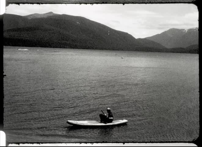 A black and white wide shot shows a person on a small boat in the middle of a large body of water surrounded by mountains under a cloudy sky