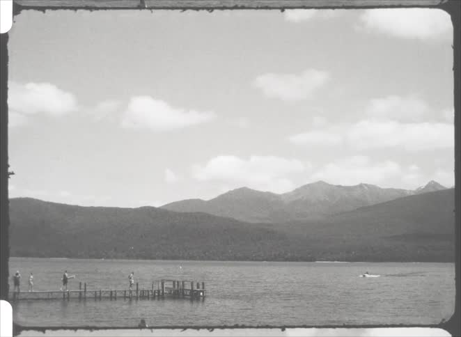 Several people walk along a long wooden pier extending into a large lake while a small motorboat cruises across the water against a backdrop of rolling hills and a prominent mountain range under a cloudy sky