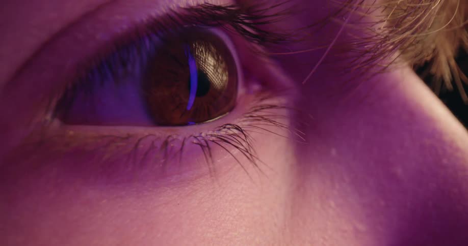 A closeup video shows a mans brown eye as he blinks capturing the detailed texture of the iris and eyelashes while colorful ambient light reflects off the surface of the eyeball against a dark background