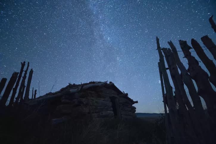 Old cabin and broken trees as the stars of the milky way and shooting stars move across the night sky