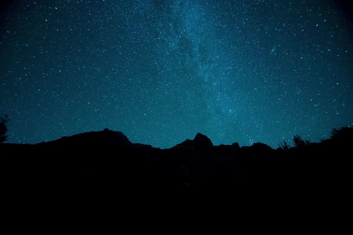 A timelapse video captures the milky way and thousands of stars rotating across a deep blue night sky over the dark silhouetted jagged peaks of a mountain range