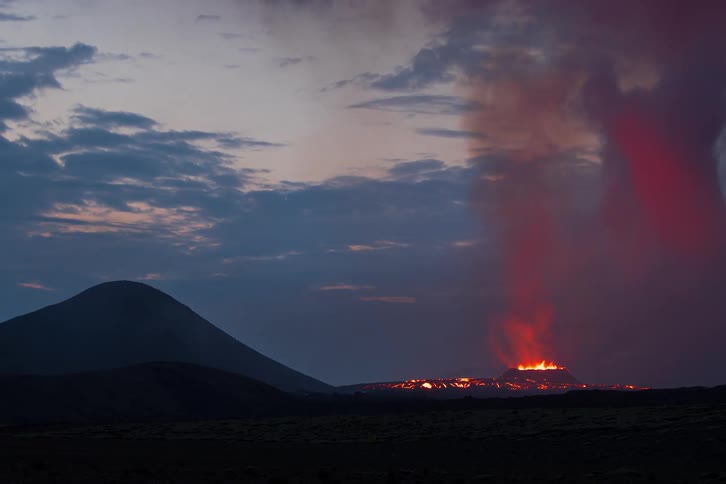 Volcanic lava sits on the lower part of the mountain at sunset glowing orange and red flames flicker on the surface and smoke rises slowly into the air
