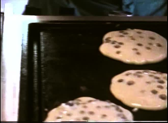Three blueberry pancakes cook on a dark griddle while a person prepares the kitchen counter nearby reaching for a paper towel next to a can of buttery cooking spray a knife and wrapped food