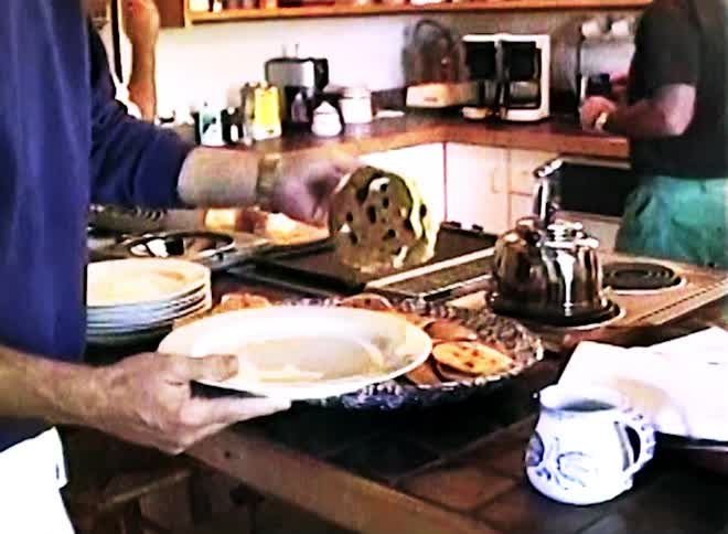 In a bright kitchen a person uses a fork to pick up several pancakes and bacon from a large serving platter and place them onto a white plate