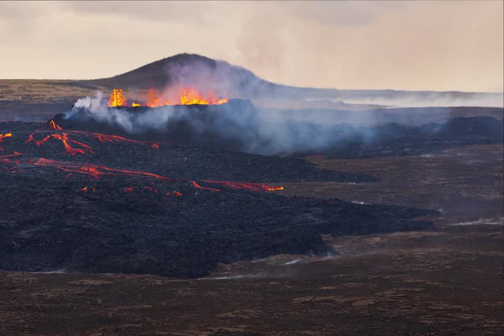 Smoke and flames emerge from an erupting volcano as molten hot lava moves across the land