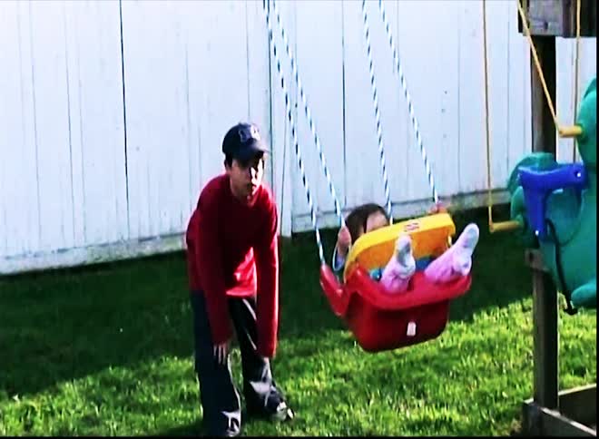 In a grassy backyard with a white fence a young boy in a red shirt and a baseball cap pushes a toddler in a red and yellow swing