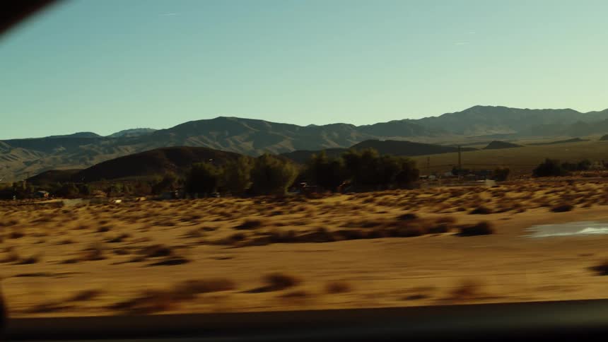 A wide shot from a moving vehicle shows a vast arid desert landscape with sparse brown vegetation and distant dark mountains under a clear bright sky