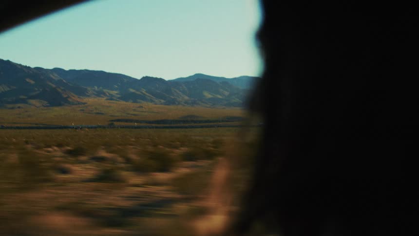 A woman sits in the passenger seat of a car driving along a countryside freeway at midday the wind coming through the window makes her hair sway gently as she gazes outside at the passing landscape
