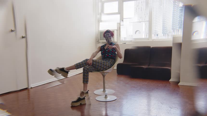 Woman seated on a swivel chair crosses her legs wearing patterned clothing and a mask in an indoor space with a window and sofa in the background