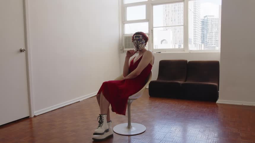 Woman seated on a swivel chair wears a red dress and a mask in an indoor space with a window and sofa in the background