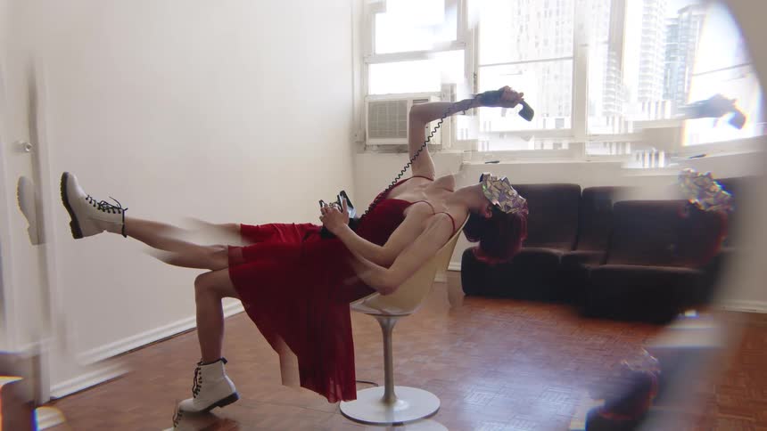 Woman reclining on a swivel chair extends her legs and holds a corded telephone in an indoor space with a window and sofa in the background