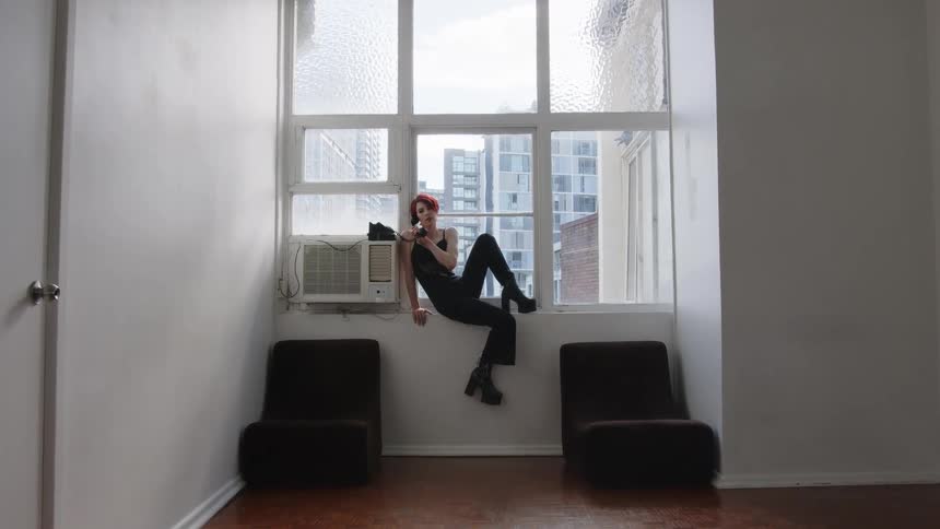 Woman sits on a windowsill holding a phone indoors with buildings visible in the background
