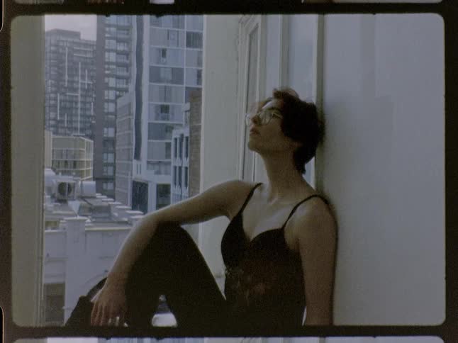 Person sitting on a window ledge touches the head with one hand while urban buildings appear in the background