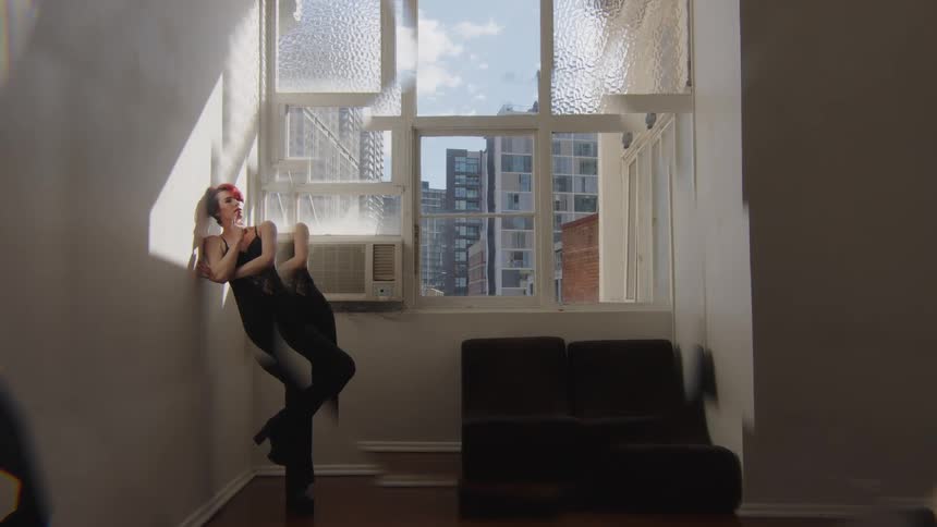 Woman leans her body backward against a wall in front of a window in an indoor space with buildings in the background