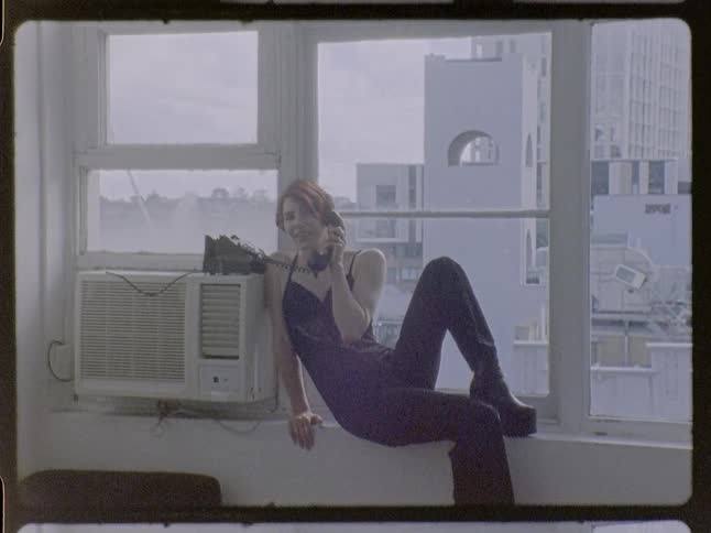 Woman sitting on a window ledge holds a corded telephone in an indoor space with buildings in the background