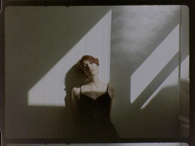 In a medium shot captured with a vintage film aesthetic a woman with short reddish brown hair in a black shirt leans against a wall in a room moving her head and body as sunlight from a nearby window creates dramatic shadows
