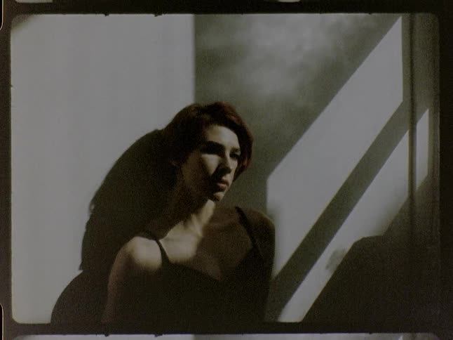 A medium shot captured with a vintage film aesthetic shows a woman with short red hair sitting in a dimly lit room as sunlight from a nearby window creates dramatic shadows across her face and the wall behind her