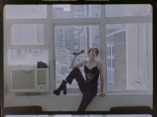 Woman sitting on a window ledge holds a corded telephone near her face in an indoor space with buildings in the background