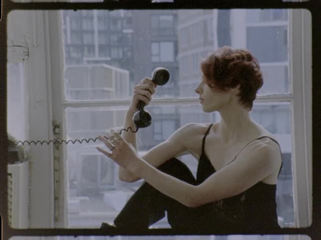 Woman sitting by a window holds a corded telephone and looks at the object indoors