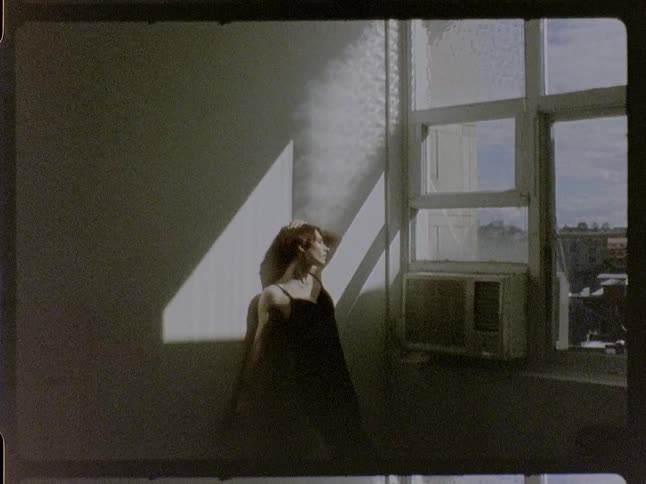 A medium shot captured with a vintage film aesthetic shows a woman with short reddishbrown hair in a black dress leaning against a wall in a room moving her arms and head as sunlight from a nearby window creates dramatic shadows