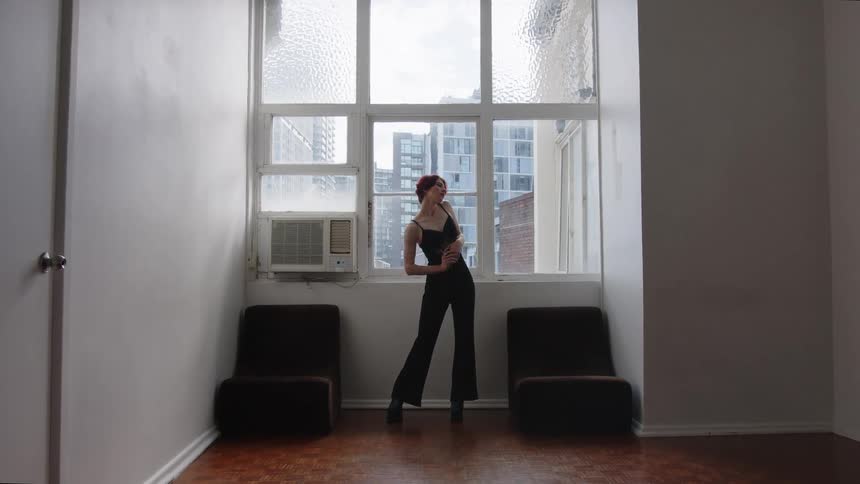 Woman standing in front of a window wears black clothing in an indoor space with buildings visible in the background