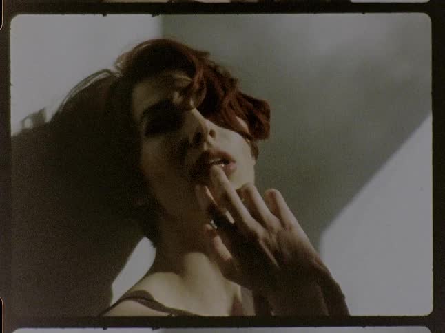 A closeup shot captured with a vintage film aesthetic shows a woman with short reddishbrown hair leaning against a wall in bright sunlight moving her hands through her hair and around her face