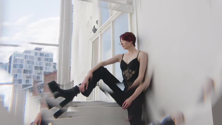 A female model with red hair sits on a balcony at midday wearing boots black trousers and a black top she poses calmly in the bright daylight