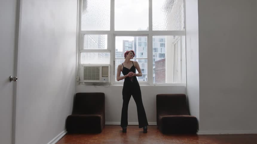 In a wide shot a woman with short red hair wearing a black sleeveless shirt and platform boots poses in an empty room with white walls and a large window overlooking the city