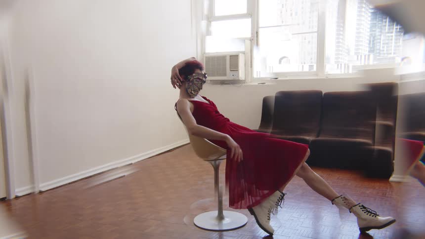 In a medium shot with a kaleidoscopic visual effect a woman wearing a red dress white boots and a reflective mosaic mask poses gracefully while reclining in a white chair within a sunlit room