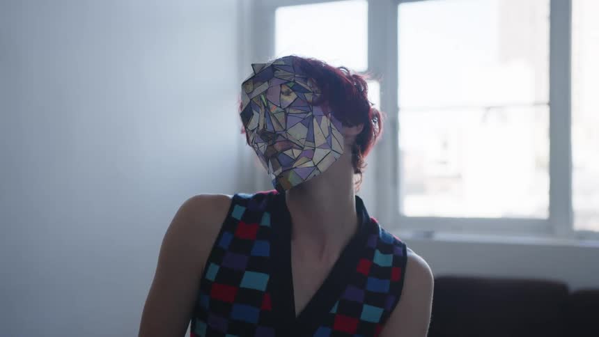 A medium shot shows a person with short red hair wearing a mask made of reflective multicolored geometric shards and a checkered vest as they slowly move their head and torso while sitting in a room with a bright window