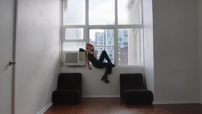 A young woman sits in an apartment windowsill while talking on a vintage phone