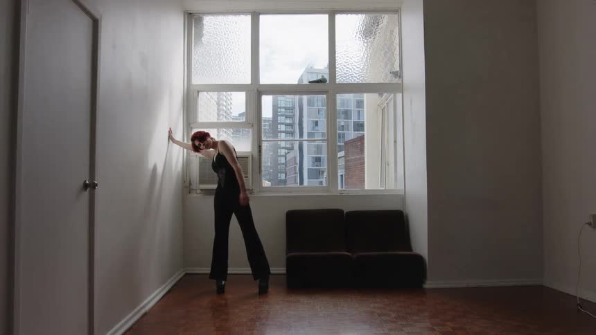 A woman rehearses slow dance movements in a sunlit minimalist room with a large city view window