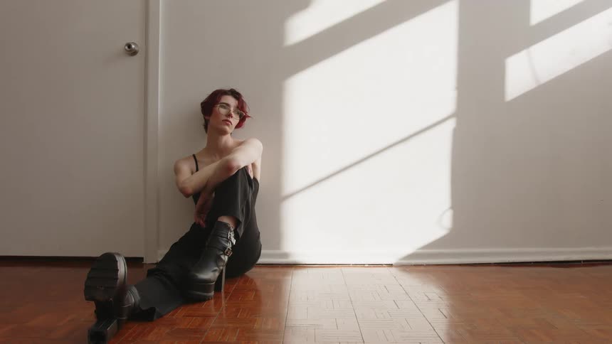 In a wide shot a person with short red hair wearing a black sleeveless shirt and platform boots sits on a wooden floor leaning against a white wall in a room where sunlight creates strong geometric shadows