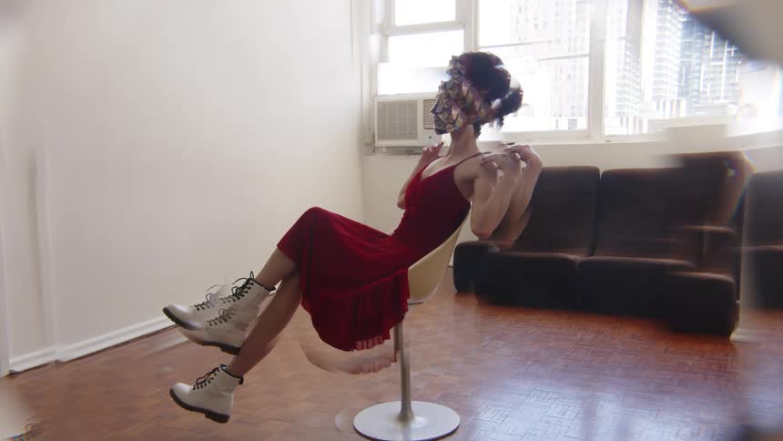 A woman wearing a red dress and white boots with a reflective mosaic mask poses gracefully while sitting in a white chair within a sunlit room with a kaleidoscopic visual effect
