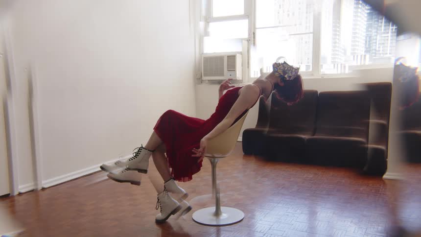 Woman reclining on a swivel chair wears a red dress and a mask with a window and sofa in the background