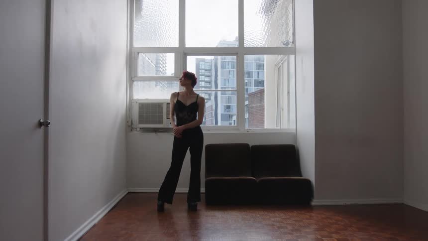 Woman standing in front of a window clasps her hands in front of her body in an indoor space with buildings in the background