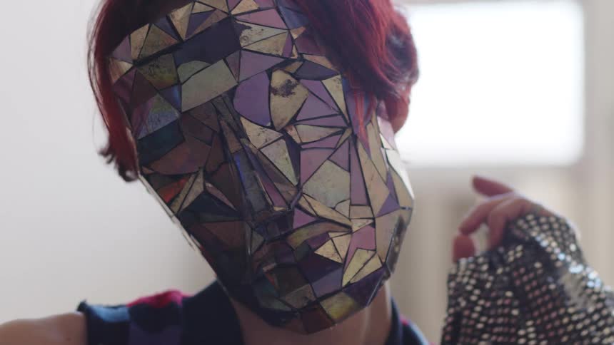 Woman wears a reflective geometric mask and raises a hand with a sparkling glove indoors