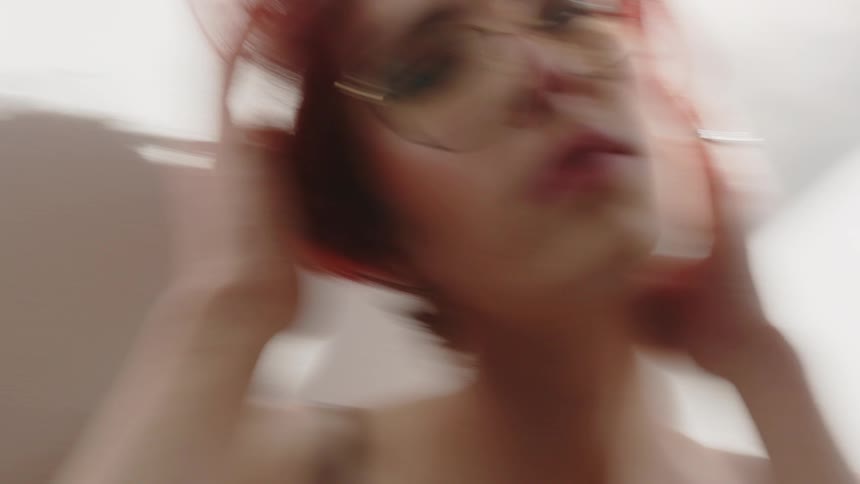 A young woman with red hair sits indoors beside a wall wearing clear glasses and a nightgown sunlight from a nearby window fills the room