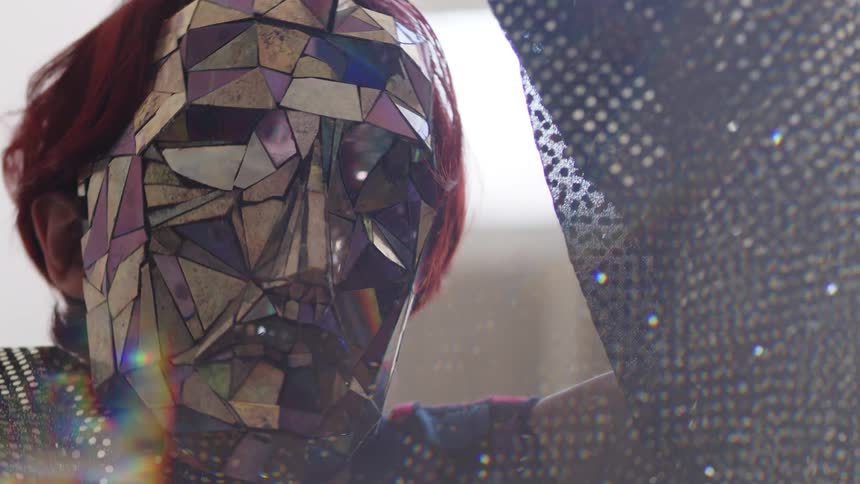 A clos up shot shows a person with red hair wearing a mask made of reflective multicolored geometric shards as they move their hands and head in front of a bright window