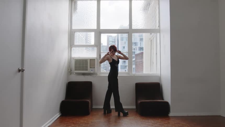 Woman standing in front of a window adjusts her mask wearing black clothing in an indoor space with buildings in the background