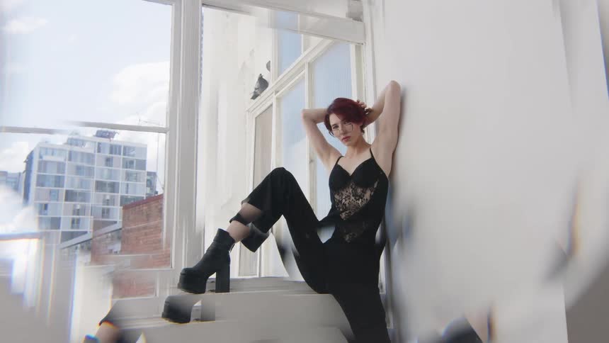 A woman with short red hair wearing a black lace top and trousers with platform boots poses against a white wall by a large window with city views while a prismatic lens effect creates shimmering light