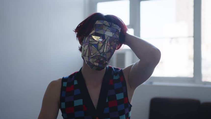 Woman wears a colorful geometric mask and touches her head in an indoor space with a window in the background