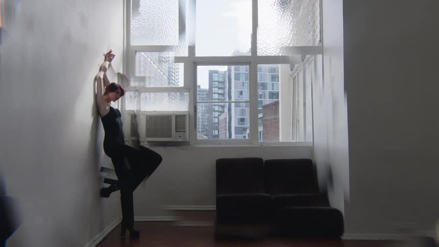 Woman supports her body against the wall with arms raised in front of a window in an indoor space with buildings in the background