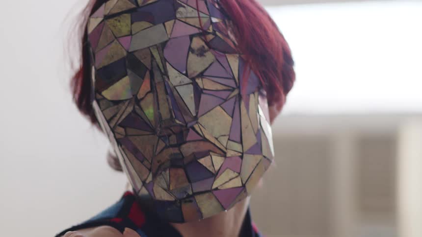A close up shot focuses on a person with short red hair wearing a mask made of reflective multicolored geometric shards as they move their hands and head in a brightly lit indoor space