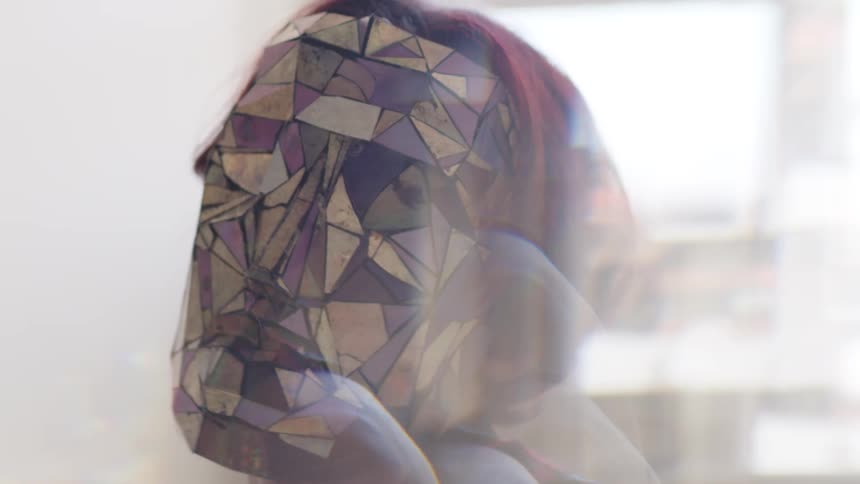 A close up shot focuses on a person with short red hair wearing a mask made of reflective multicolored geometric shards as they move their hands and head in a brightly lit indoor space